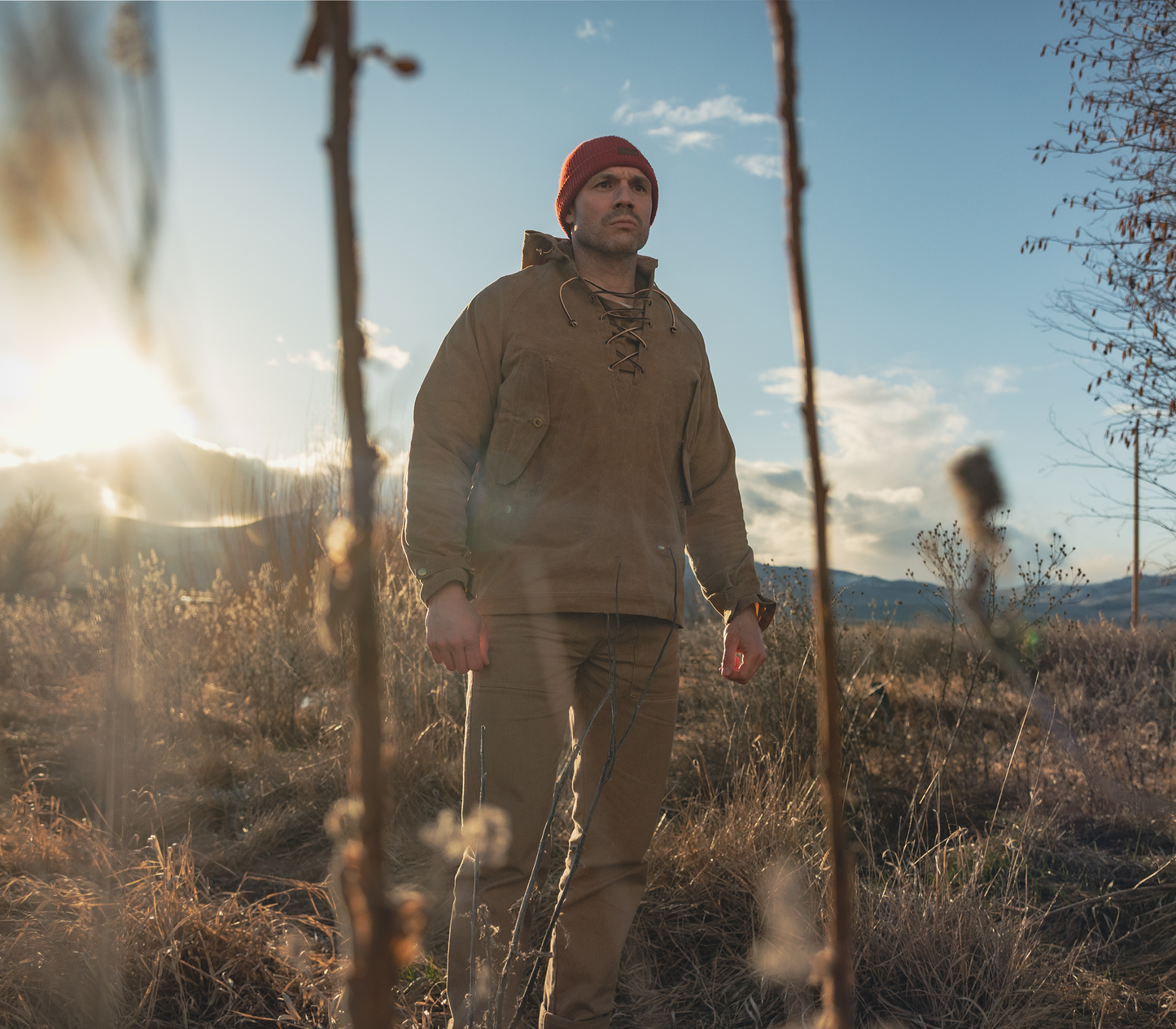 A man wearing a tan outdoor work jacket and matching pants with a red beanie stands outdoors in a field of dry grass under bright sunlight.