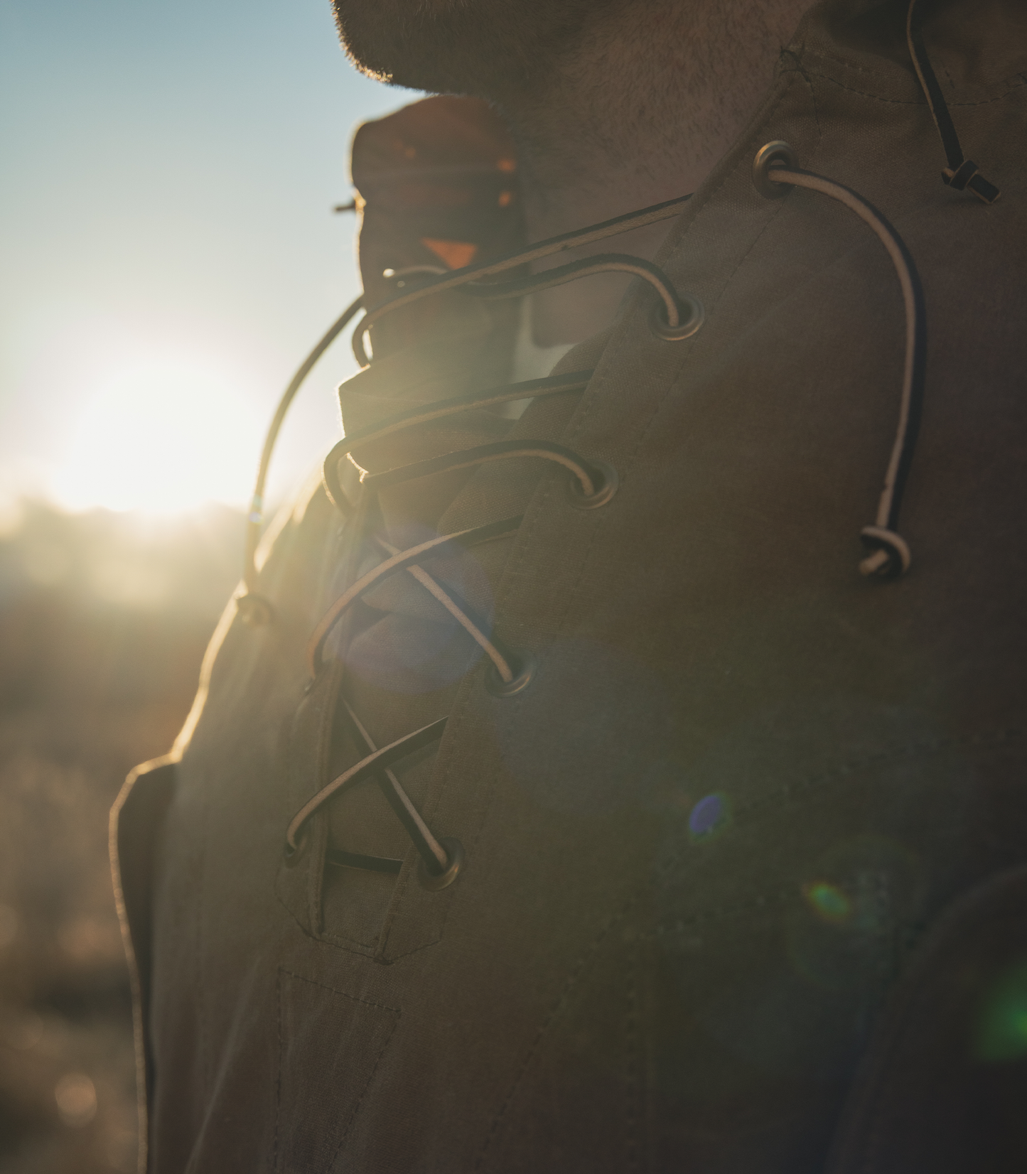 Close-up of a person outdoors wearing a dark brown or olive lace-up jacket, with the sun low in the background causing strong backlighting and lens flare.