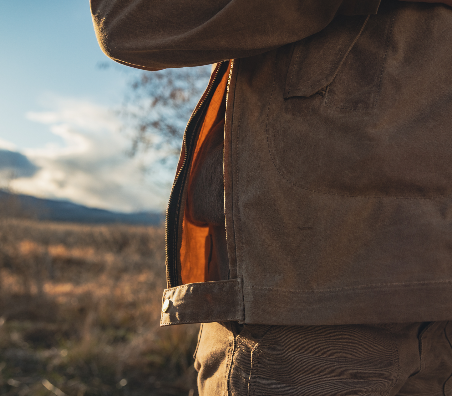 A close-up of a brown waxed canvas bag with an orange-brown interior lining, shown outdoors with a blurred mountain and sky background.